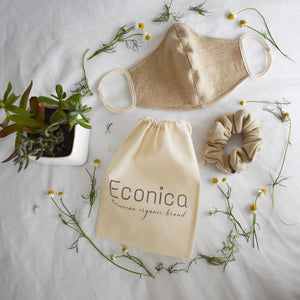 Una mascarilla de tela color marrón claro, una bolsa de tela que dice "Ecónica - Peruvian organic brand", un scrunchy marrón claro, una planta pequeña en una maceta y flores de manzanilla sobre una superficie blanca. 